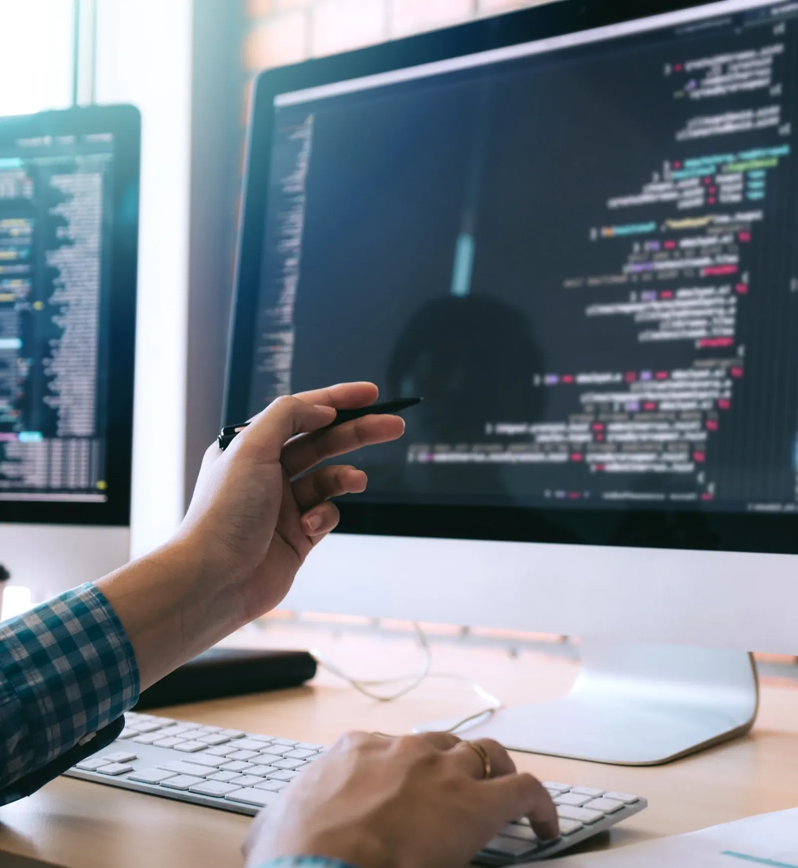 Hand vor einem Bildschirm mit Quelltext, ein zweiter Monitor daneben